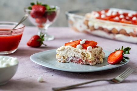Italian traditional dessert tiramisu with strawberries. One piece of cake on a plate closeup, whole cake and strawberry juice backside, fork and knife, rose and grey concrete background